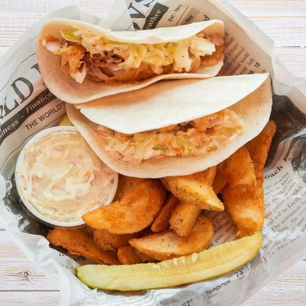 Two fish tacos with coleslaw, a side of potato wedges, a pickle spear, and dipping sauce.