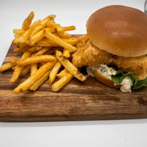 Crispy fried fish sandwich with tartar sauce and lettuce, served with a side of seasoned fries.