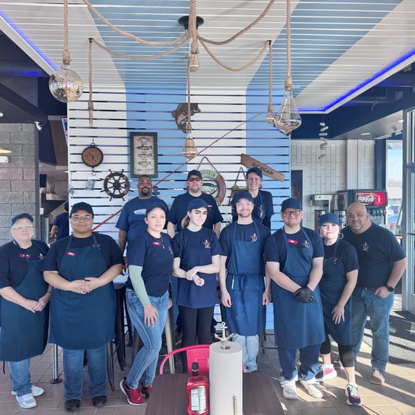 Eleven restaurant staff members in blue uniforms and aprons pose together in a brightly lit dining area.
