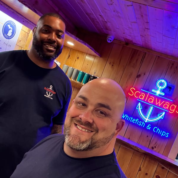 Two smiling men in Scalawags shirts stand in front of a neon sign for Scalawags Whitefish & Chips.
