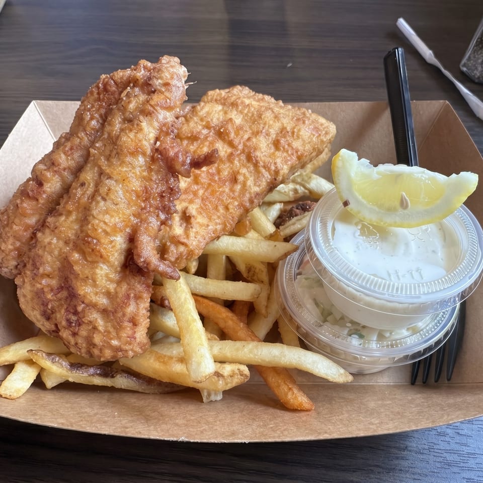 Golden-brown fish and chips in a cardboard tray with tartar sauce and a lemon wedge.