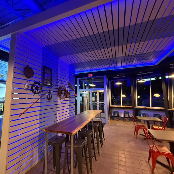 Restaurant interior with blue lighting, nautical decor, high-top tables, and red chairs.