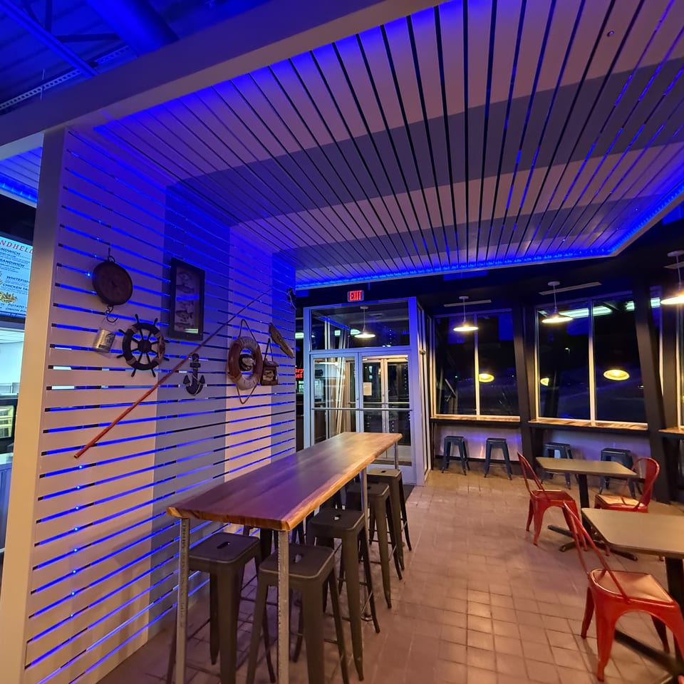 Restaurant interior with blue lighting, nautical decor, high-top tables, and red chairs.