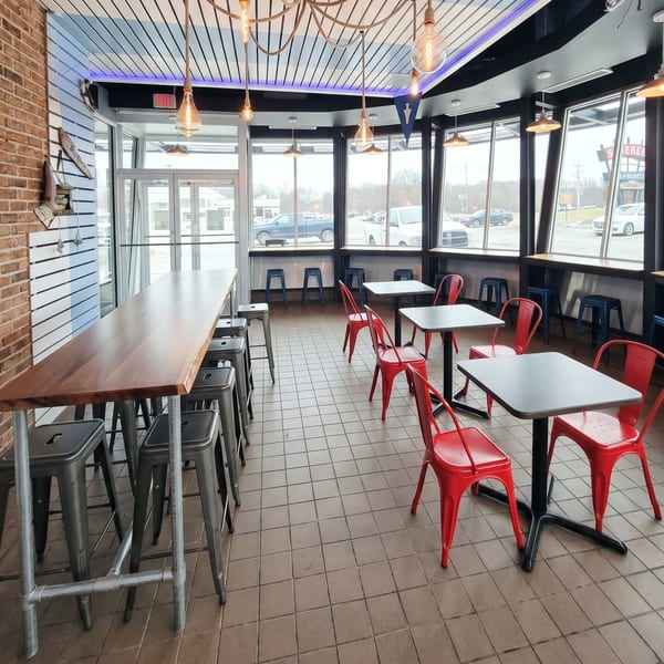 Restaurant interior with a long wooden bar, red chairs, and large windows overlooking a parking lot.
